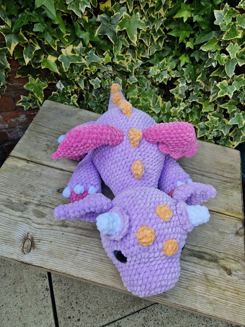 Pu&ograve; includere: Un giocattolo di peluche di drago di colore viola lavorato a uncinetto con ali rosa, macchie arancioni e corna bianche. Il drago &egrave; sdraiato su una superficie di legno.