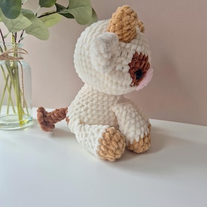 May include: A crocheted plush toy in the shape of a cow. The cow is white with brown spots and has a brown tail. The cow is sitting on a white surface.