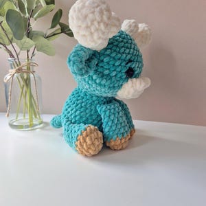 Pu&ograve; includere: Un giocattolo di peluche color turchese lavorato a uncinetto a forma di dinosauro triceratopo con corna bianche e piedi marroni.
