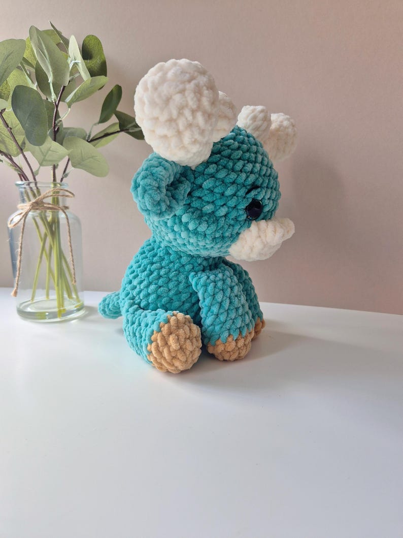 Pu&ograve; includere: Un giocattolo di peluche color turchese lavorato a uncinetto a forma di dinosauro triceratopo con corna bianche e piedi marroni.