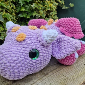 Pu&ograve; includere: Un giocattolo di peluche a forma di drago, lavorato a uncinetto, di colore viola e rosa con dettagli arancioni e bianchi. Il drago ha un occhio verde scintillante e un corno bianco.