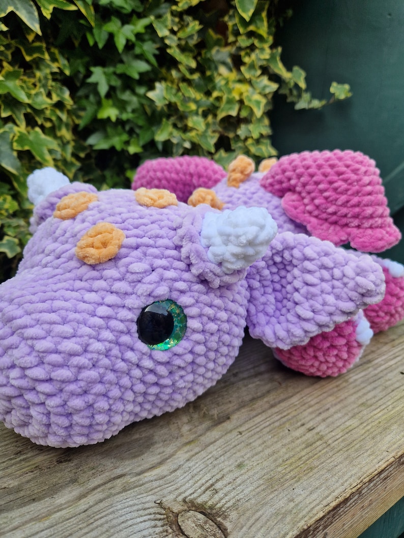 Pu&ograve; includere: Un giocattolo di peluche a forma di drago, lavorato a uncinetto, di colore viola e rosa con dettagli arancioni e bianchi. Il drago ha un occhio verde scintillante e un corno bianco.