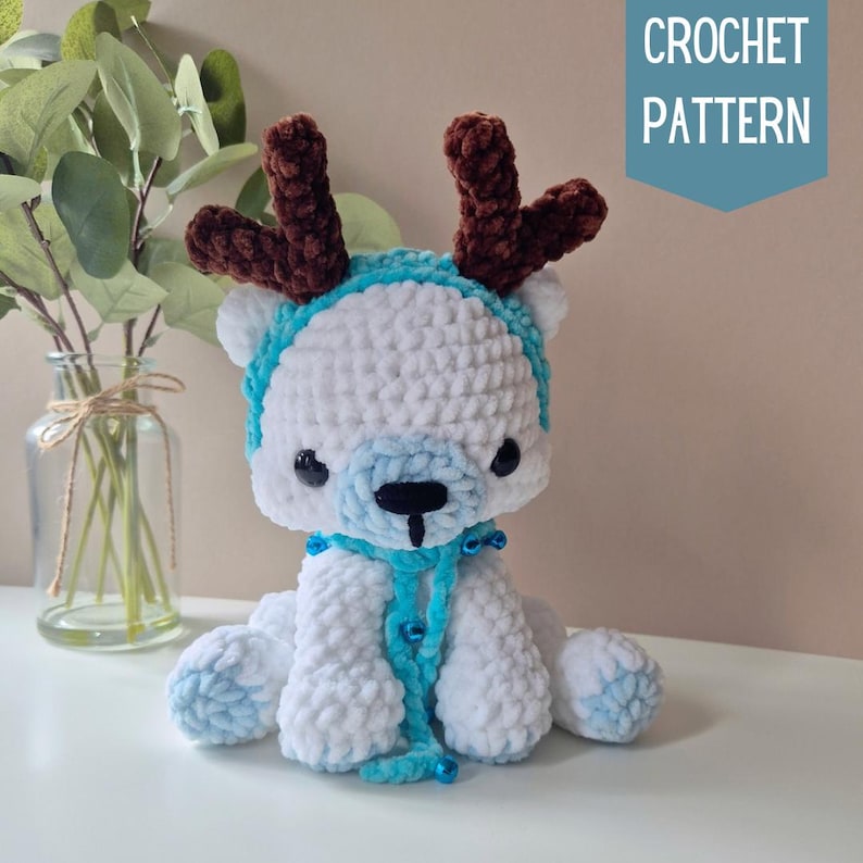 May include: White crocheted reindeer with blue and brown accents. The reindeer is wearing a blue scarf and has brown antlers. CROCHET PATTERN text is visible in the top right corner of the image.