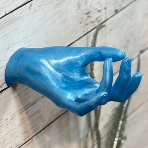Könnte beinhalten: Eine blaue Handskulptur, montiert an einer weißen Holzwand. Die Hand ist in einer Geste, mit leicht gekrümmten Fingern. Die Skulptur hat einen metallischen Glanz, und die Wand hat ein rustikales, verwittertes Aussehen.
