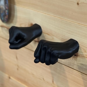 May include: Two matte black wall hooks shaped like human fists, mounted horizontally on a light-colored wooden plank wall. Each hook features a detailed, realistic hand design with fingers curled into a tight fist, extending outward from the surface.