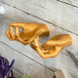 May include: Gold-coloured, wall-mounted hand sculpture with open palms, mounted on a light wood-panelled wall. The sculpture is a decorative accent piece. Other decorative items are visible in the background.