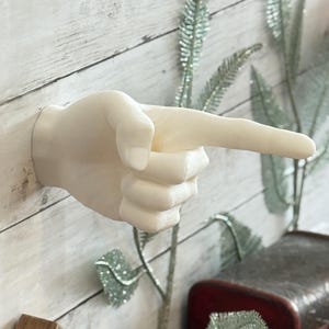 Könnte beinhalten: Eine mattweiße Wandskulptur in Form einer menschlichen Hand, bei der der Zeigefinger in einer zeigenden Geste ausgestreckt ist. Die Hand ist horizontal positioniert, wobei die Handfläche nach unten zeigt und der Daumen an den Fingern anliegt.