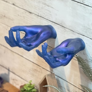 Könnte beinhalten: Zwei blaue, offene Handskulpturen an einer Holzwand befestigt. Die Hände haben ein metallisches, irisierendes Finish mit Blau- und Lilatönen. Die Finger sind leicht gekrümmt. Dekorative Kunst.