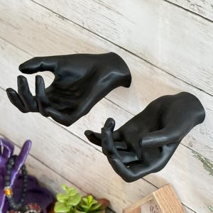 Könnte beinhalten: Zwei schwarze Handskulpturen, montiert an einer hellen Holzpaneelwand. Die Hände sind in einer Geste des Anbietens oder Empfangens, mit leicht gekrümmten Fingern. Die Skulpturen sind der Mittelpunkt, mit anderen dekorativen Elementen im Hintergrund.