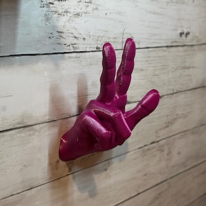 May include: A bright pink 3D printed hand sculpture making the peace sign. The hand is mounted on a white wooden wall.
