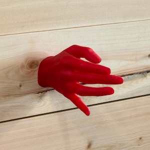 May include: A bright red, hand-shaped wall hook mounted on a light-colored wooden surface. The hand is positioned with fingers extended, creating a functional hook for hanging items. The design features a smooth, matte finish and realistic details.