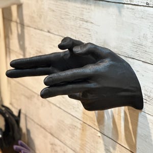 Könnte beinhalten: Schwarze Handskulptur, die eine Geste ausführt, an einer hellen Holzwand befestigt. Der Zeige- und Mittelfinger sind ausgestreckt, die anderen Finger sind gefaltet.