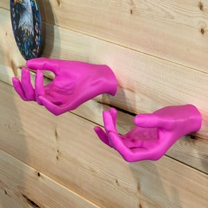 May include: Two bright pink, open-palm hand sculptures mounted to a wall. Each hand is positioned with fingers slightly curled upward, creating a cupped shape. The smooth, matte finish of the hands contrasts with the light-colored wood grain wall.