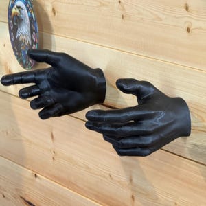 Könnte beinhalten: Zwei mattschwarze, an der Wand montierte Handskulpturen, die nach außen ragen. Jede Hand weist realistische anatomische Details auf, einschließlich definierter Finger, Knöchel und Handlinien, die so positioniert sind, als würden sie hervorragen.