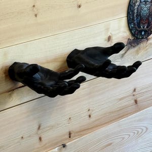 May include: Two matte black wall-mounted sculptures shaped like human hands, positioned side-by-side with palms facing upward. Each hand features realistic finger articulation and wrist detail, extending outward from the flat mounting surface.