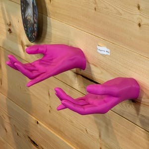 Könnte beinhalten: Zwei leuchtend magentafarbene, matt lackierte Wandskulpturen in Form von Händen sind nebeneinander angebracht. Jede Hand ist geöffnet, die Handflächen zeigen nach oben und ragen aus der Wand heraus. Ein kleines weißes Etikett mit der Aufschrift Magenta, Matte ist zu sehen.