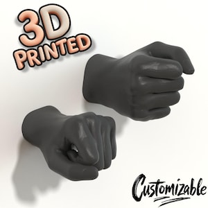Könnte beinhalten: Zwei dunkelgraue, 3D-gedruckte Wandhaken in Faustform. Der Text 3D PRINTED erscheint in der oberen linken Ecke und das Wort Customizable ist in schwarzer Schreibschrift in der unteren rechten Ecke zu sehen. Die Fäuste wirken wie aus der Wand kommend.