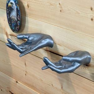 May include: Two silver-colored, realistic hand sculptures mounted to a wall. Each hand is positioned with the palm facing upward, fingers slightly curled, and the thumb extended. The surface of the hands has a smooth, metallic sheen.