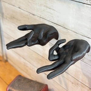 Può includere: Due sculture da parete nero opaco a forma di mani umane. Ogni mano è posizionata con le dita estese in un gesto specifico, sembrando emergere dalla superficie della parete. Le sculture presentano una finitura scura e liscia.