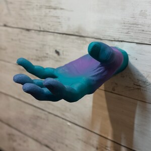 Könnte beinhalten: Eine Wandskulptur in Form einer Hand in Blau-, Türkis- und Lilatönen. Die Hand ist ausgestreckt, als ob sie etwas anbietet oder empfängt. Die Skulptur ist an einer hellen Wand aus Holzbrettern befestigt.
