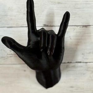 Könnte beinhalten: Eine mattschwarze Wandskulptur einer Hand, die das Ich-liebe-dich-Zeichen in amerikanischer Gebärdensprache zeigt. Die Hand ist so positioniert, dass der Daumen abgespreizt, Zeige- und kleiner Finger erhoben und Mittel- und Ringfinger eingeknickt sind.