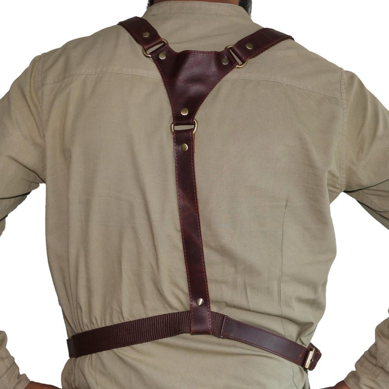 May include: Brown leather camera strap with adjustable straps and brass hardware. The strap is worn over a person's shoulders and around their waist.