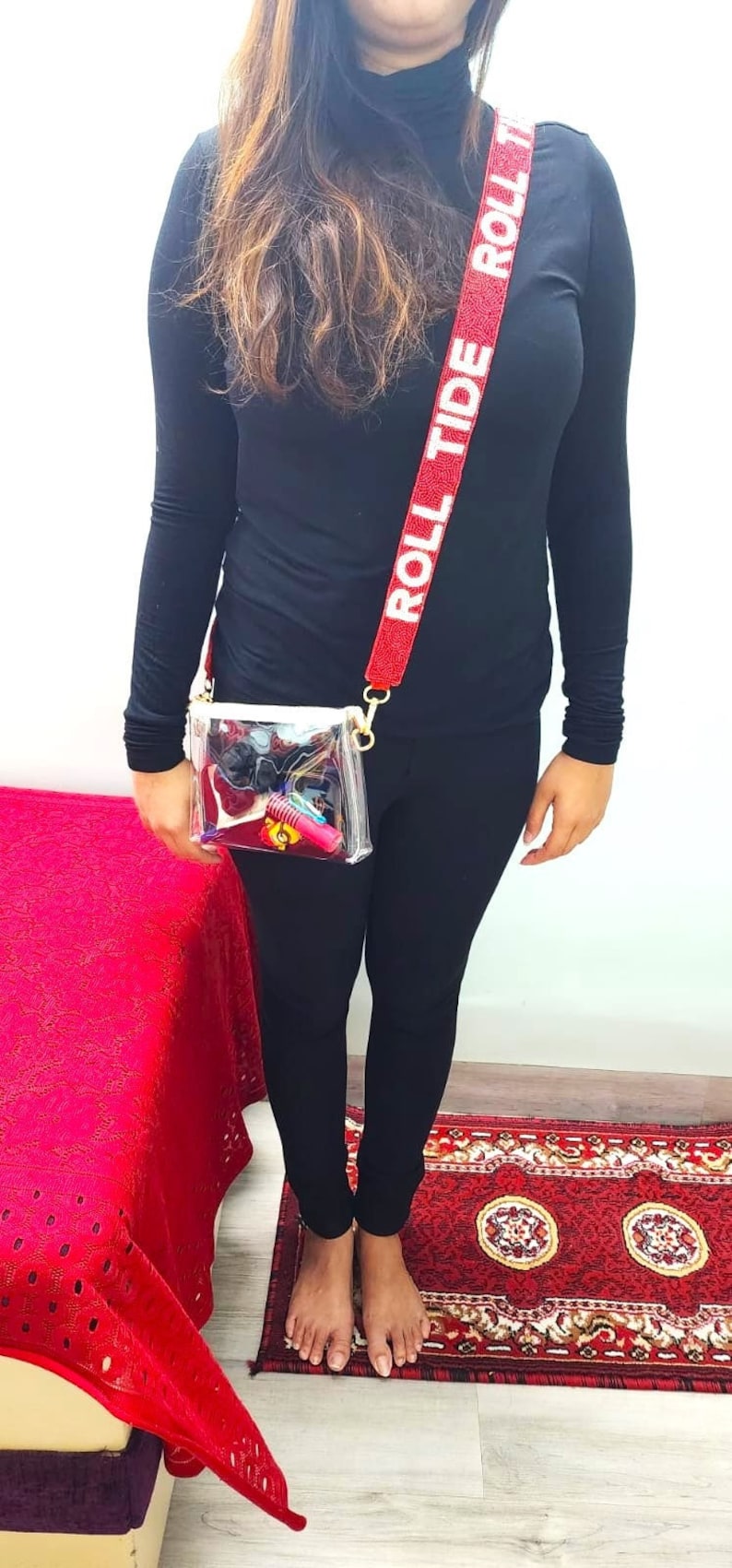 May include: A clear plastic crossbody bag with a red and white strap that says "Roll Tide". The bag is worn by a person wearing black pants and a black turtleneck shirt.
