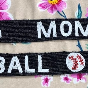 May include: Two black fabric wristbands with white text. The top band says "Boxing Momma" with a red boxing glove icon on each end. The bottom band says "Play Ball" with a baseball icon on each end.