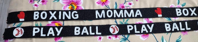 May include: Two black fabric wristbands with white text. The top band says "Boxing Momma" with a red boxing glove icon on each end. The bottom band says "Play Ball" with a baseball icon on each end.