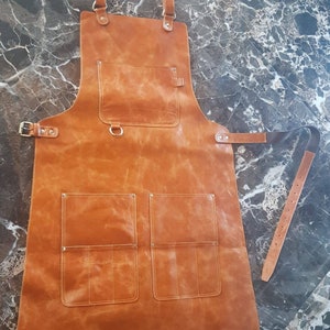 May include: A brown leather apron with multiple pockets and adjustable straps. The apron has a D-ring on the front and a belt loop on the side.