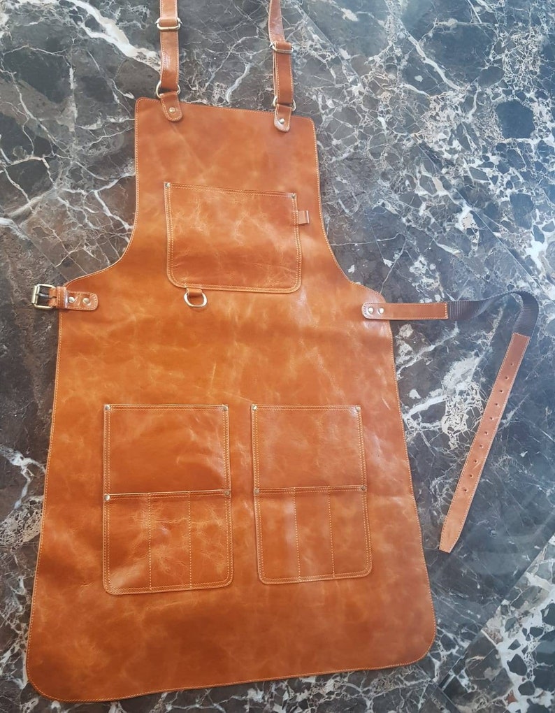 May include: A brown leather apron with multiple pockets and adjustable straps. The apron has a D-ring on the front and a belt loop on the side.