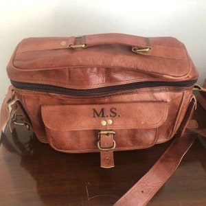 May include: A brown leather travel bag with a top handle and a shoulder strap. The bag has a front pocket with a buckle closure and the initials "M.S." embroidered on it.