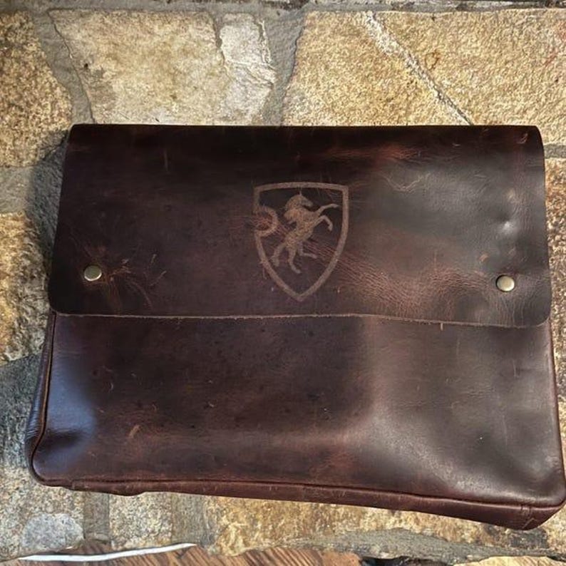 May include: A brown leather laptop sleeve with a horse head logo in a shield shape.