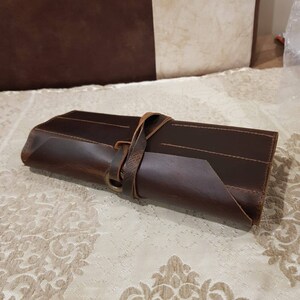 May include: A brown leather roll-up case with a strap and buckle closure. The case is designed to hold and protect multiple items, such as knives or tools.