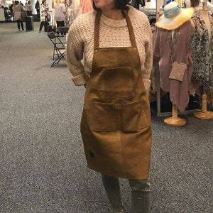 May include: A brown leather apron with two large pockets and a smaller pocket on the front. The apron is worn over a white sweater.