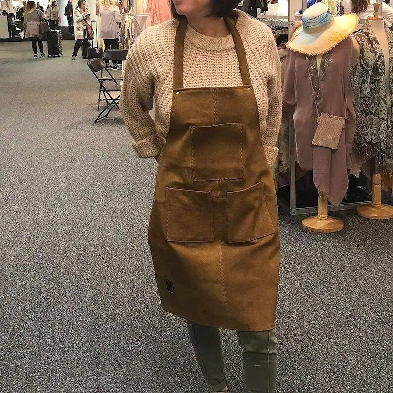 May include: A brown leather apron with two large pockets and a smaller pocket on the front. The apron is worn over a white sweater.