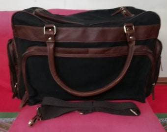 May include: Black canvas and brown leather duffel bag with a shoulder strap. The bag has two top handles and a zipper closure.