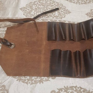 May include: A brown leather roll-up case with multiple slots for holding tools. The case has a strap for carrying.