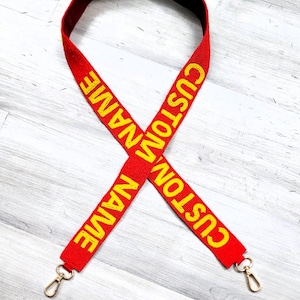 May include: Red adjustable strap with gold metal clasps. The strap is embroidered with the text "CUSTOM NAME" in yellow.