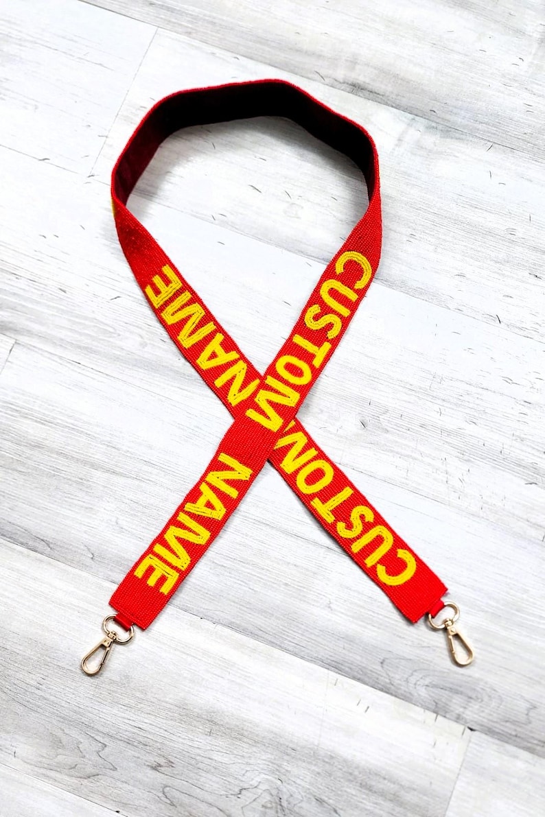 May include: Red adjustable strap with gold metal clasps. The strap is embroidered with the text "CUSTOM NAME" in yellow.