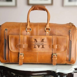 May include: A tan leather duffel bag featuring two top handles, two vertical zippered side pockets, and a front flap pocket secured with two buckled straps. The front pocket is engraved with a large letter M, the name Marcus, and a horizontal line.