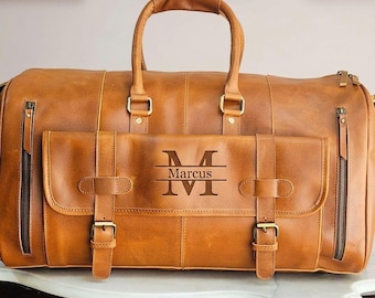 May include: A tan leather duffel bag featuring two top handles, two vertical zippered side pockets, and a front flap pocket secured with two buckled straps. The front pocket is engraved with a large letter M, the name Marcus, and a horizontal line.