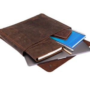May include: A brown leather laptop bag with a flap closure and a zippered pocket. The bag is open and contains a laptop, a notebook, and a pen.