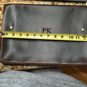 May include: A brown leather laptop sleeve with the initials "PK" embossed on the front. The sleeve is measured with a tape measure, showing it is 12 inches wide.