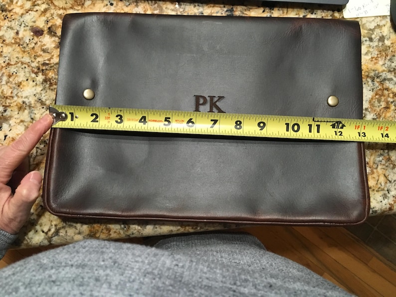 May include: A brown leather laptop sleeve with the initials "PK" embossed on the front. The sleeve is measured with a tape measure, showing it is 12 inches wide.