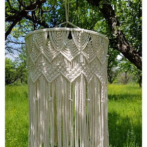 May include: A white macrame lampshade hanging from a tree branch. The lampshade is made of a thick, natural fiber rope and has a geometric pattern. The lampshade is hanging in a garden setting with green grass and trees in the background.
