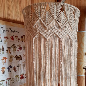 May include: A beige macrame lampshade with a geometric pattern and long fringe. The lampshade is hanging from a ceiling fixture.
