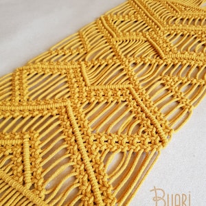 Puede incluir: Un tapiz de macramé de color amarillo mostaza con un patrón geométrico de diamantes.