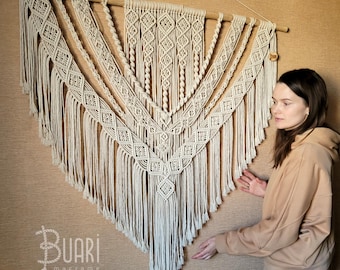 May include: A large, white macrame wall hanging with a geometric pattern and fringe. The hanging is made with natural cotton cord and is approximately 3 feet wide and 4 feet tall. The hanging is displayed on a beige wall with the text "Buari macrame" at the bottom.