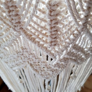 May include: A close-up of a white macrame wall hanging with a geometric pattern and fringe. The macrame is made with natural cotton cord.